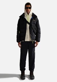 Giacca a vento nera con cappuccio, con accenti verde neon e una toppa blu. Abbinata a una felpa beige e pantaloni cargo neri. Stivali neri.