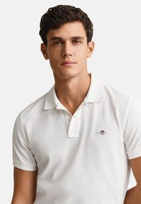 Young man with short curly brown hair wearing a white polo shirt with a small embroidered logo on the chest, neutral background.