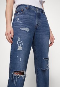 Jeans de mezclilla azul descoloridos con un corte holgado, que presentan detalles rasgados en las rodillas y el muslo, y herrajes de cobre en la cintura.