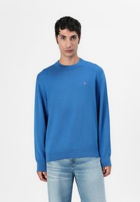 Maglione blu in cotone con collo rotondo, maniche lunghe e orlo a costine, caratterizzato da un piccolo logo rosso sul petto. Texture liscia, design casual.