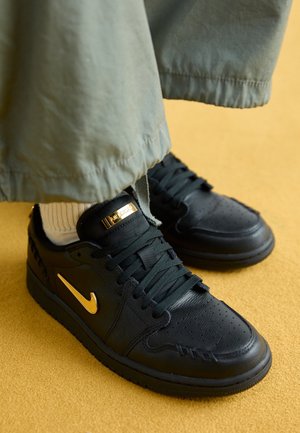 Zwarte leren sneakers met gouden Nike-swoosh en label, gedragen met beige sokken en losse olijfgroene broek op een mosterdkleurige vloer.