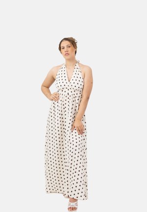 Woman wearing a white halter neck maxi dress with black polka dots and white strappy sandals, standing with one hand on her hip.