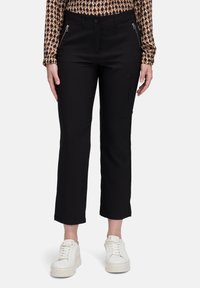 Vrouw in zwarte cropped straight-leg broek met ritszakken en witte platform sneakers, staand voor een effen achtergrond.