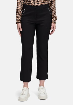 Donna che indossa pantaloni neri a gamba dritta cropped con tasche con cerniera e sneakers bianche con piattaforma, in piedi contro uno sfondo neutro.