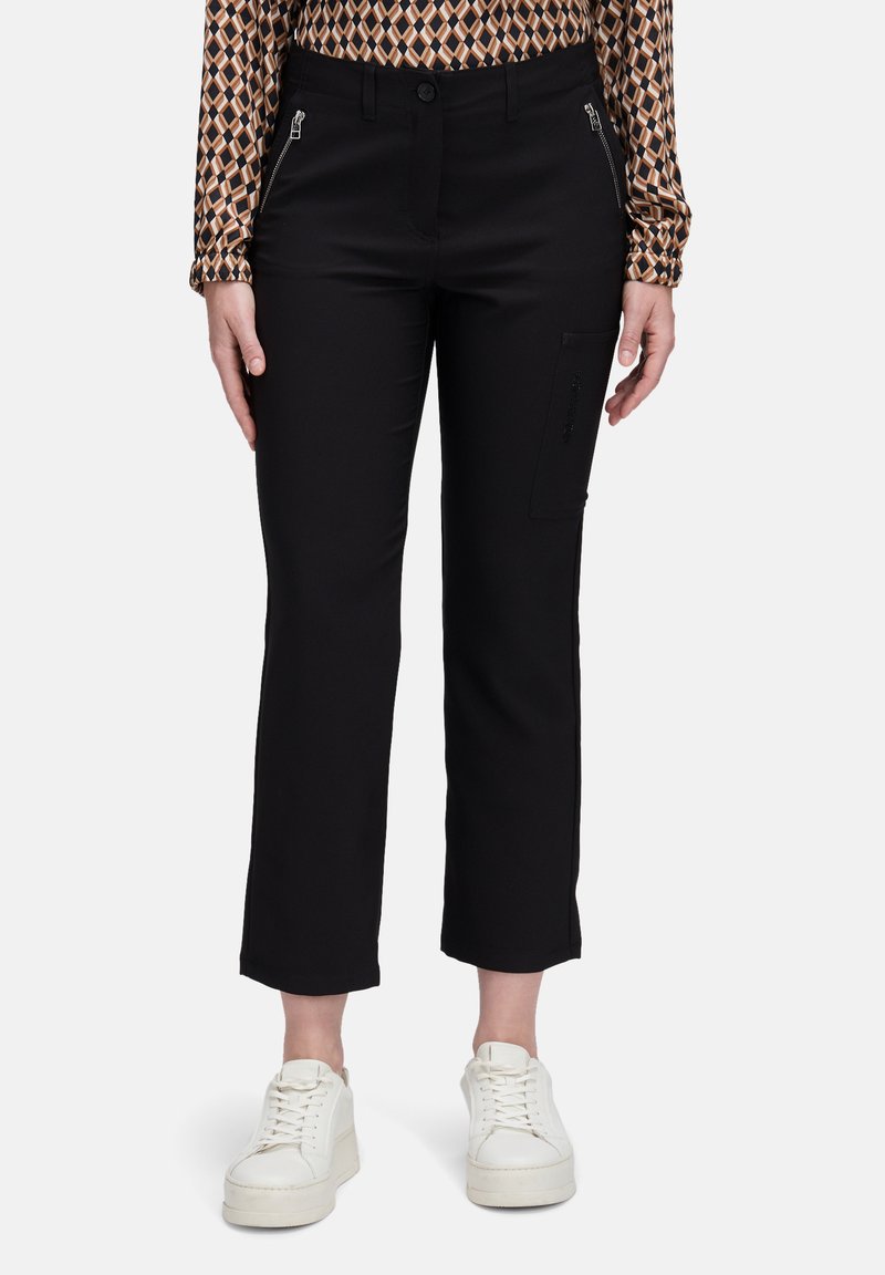 Vrouw in zwarte cropped straight-leg broek met ritszakken en witte platform sneakers, staand voor een effen achtergrond.