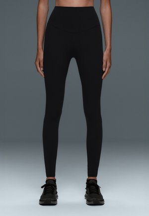 Personne portant des leggings noirs taille haute et des chaussures de sport noires, debout devant un fond gris uni.