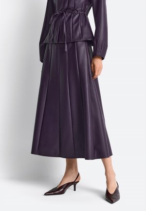 Žena v purpurovej plisovanej koženej midi sukni a zodpovedajúcom páse s dlhými rukávmi, v kombinácii s tmavými špicatými slingback opätkami.