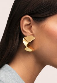 Pendiente llamativo de color dorado con un diseño escultórico de forma curva y geométrica y acabado brillante.