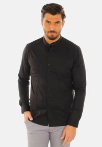 Camicia nera a maniche lunghe con colletto a band, chiusura frontale con bottoni e una texture liscia, indossata con pantaloni grigi.