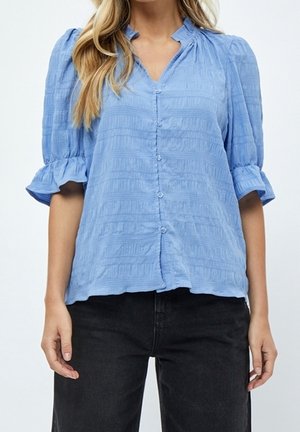 Femme portant un chemisier texturé bleu clair boutonné, avec des manches bouffantes et des poignets volantés, associé à un pantalon noir.