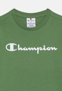 Zöld pamut póló, az elején fehér "Champion" logóval. Klasszikus kereknyakú kialakítás, címke varrva a gallér belső részébe.