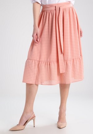 Femme portant une jupe midi à carreaux rose avec une ceinture nouée et un ourlet à volants, associée à des escarpins slingback beiges à bout pointu.