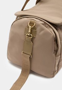 Borsa a mano in nylon beige con forma arrotondata, accessori in oro e manico intrecciato testurizzato con lettere in rilievo. Chiusura con zip in cima.