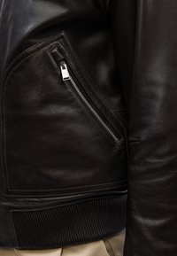 Giubbotto bomber in pelle marrone scuro con tasca con zip, orlo a coste e superficie liscia. Presenta dettagli di cucitura a contrasto.