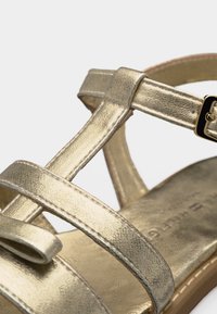 Χρυσό μεταλλικό σανδάλι με λουράκια, ρυθμιζόμενο κουμπωτό και επίπεδη σόλα, που δείχνει κοντινές λεπτομέρειες των λουριών και των ραφών.