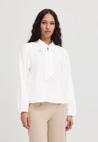 Blouse blanche avec un détail de nœud au cou, manches longues et coupe décontractée. Tissu léger avec une surface légèrement texturée.