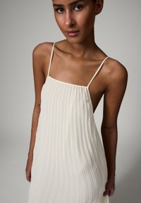 Jeune femme portant une robe blanche plissée sans manches avec fines bretelles, debout devant un fond uni gris clair.