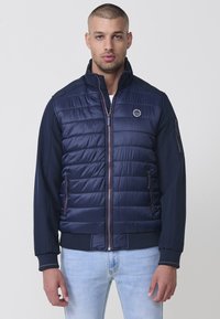 Námornícka puffer bunda s hladkým, prešívaným przedom a pletenými rukávmi. Obsahuje zips, bočné vrecká a malé logo na hrudi.