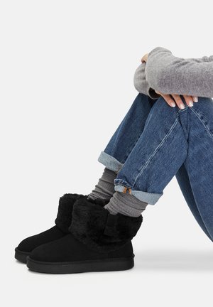 Persona seduta con le braccia attorno alle ginocchia, indossando jeans blu arrotolati, calzini grigi, un maglione grigio e stivali alla caviglia foderati in pelliccia nera.