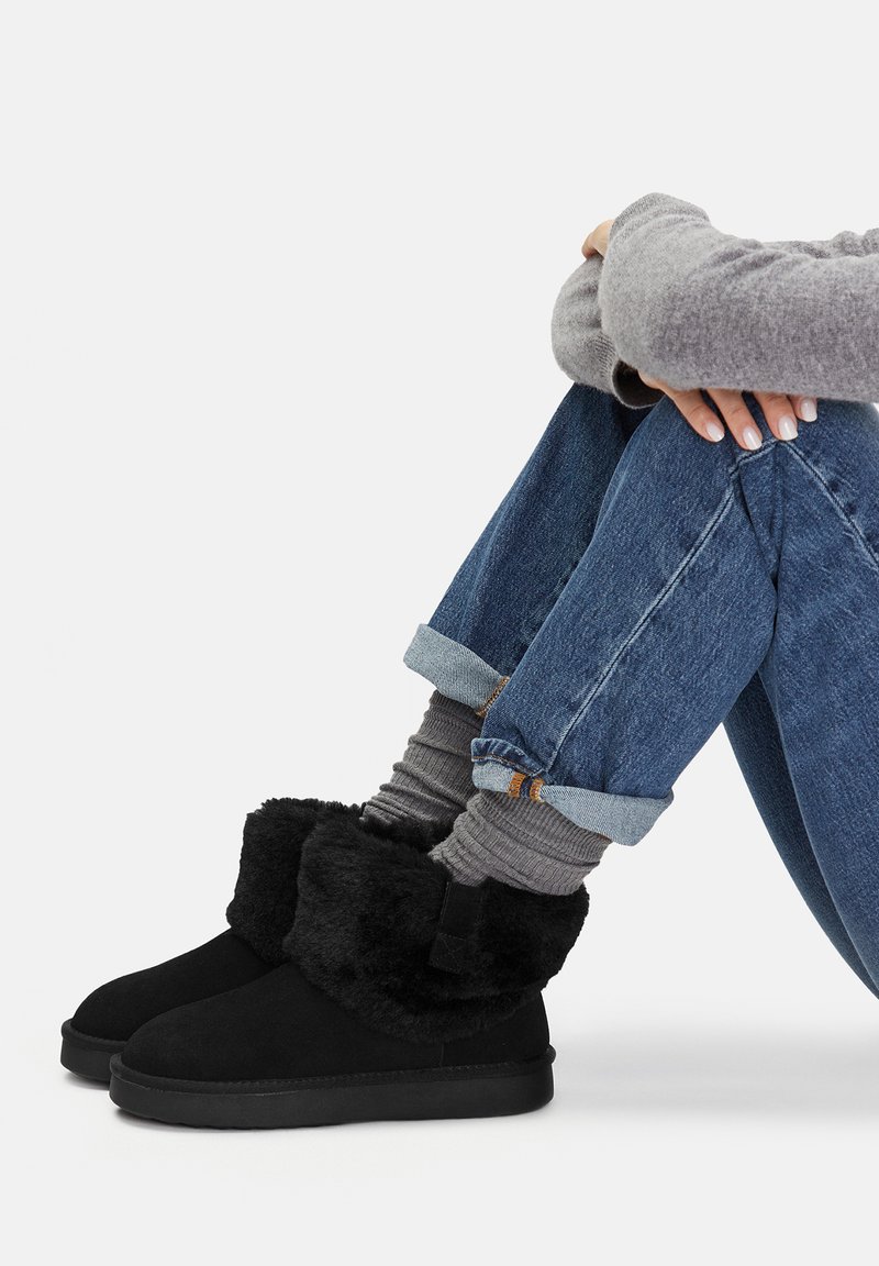 Persoană așezată cu brațele înfășurate în jurul genunchilor, purtând blugi rulați, șosete gri, pulover gri și cizme negre cu guler de blană.