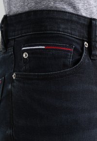 Mörkblå denimjeans med en närbild av fickan, som har en röd och vit randig accent samt silverdetaljer.