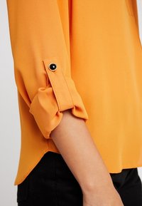 Orange blouse with 3/4 sleeves, featuring rolled cuffs secured by a black button detail. Made of smooth, lightweight fabric.