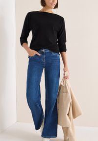 Maglione nero a maniche lunghe, jeans blu a gamba larga, sneakers bianche, mentre tiene in mano una giacca in corduroy beige chiaro. Texture liscia e vestibilità casual.