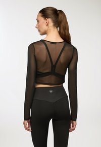 Czarny krótki top z długim rękawem wykonany z przezroczystego materiału, z konstrucją w stylu racerback. Zestawiony z czarnymi legginsami o wysokim stanie z logo na plecach.