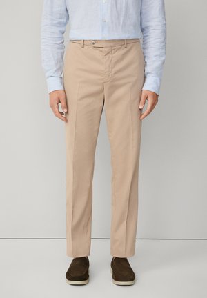 Mann trägt beige maßgeschneiderte Hose, hellblau gestreiftes Hemd mit Knöpfen und dunkelbraune Slipper, steht an einer schlichten Wand.