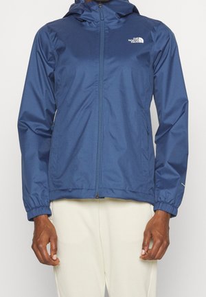 Chaqueta de lluvia azul con capucha y cremallera, puños elásticos y logo blanco en el pecho, combinada con pantalones de color claro.