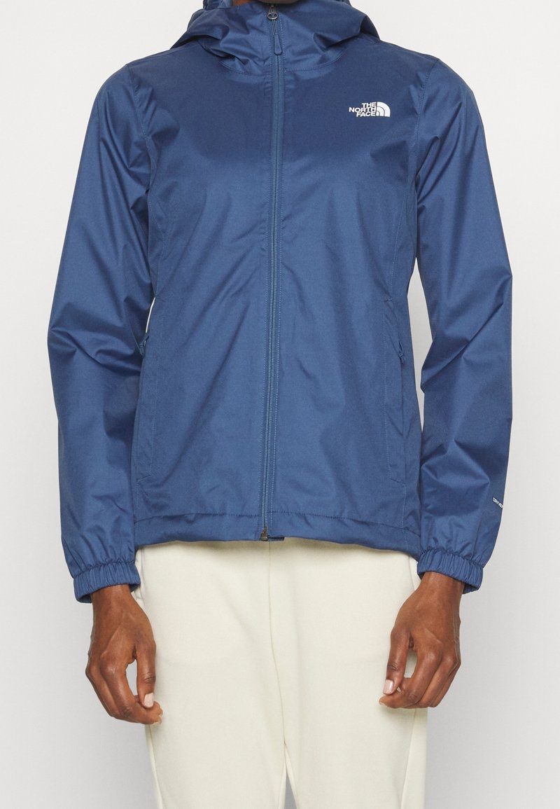 Veste de pluie bleue à capuche avec fermeture éclair, poignets élastiques et logo blanc sur la poitrine, portée avec un pantalon de couleur claire.