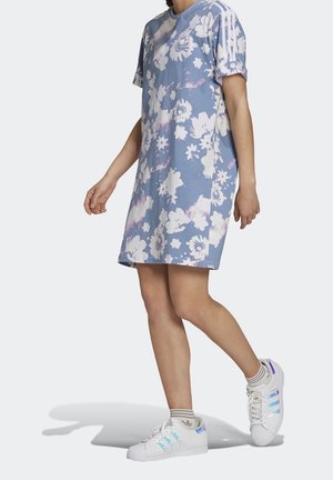 Femme portant une robe Adidas fleurie bleue et des baskets blanches avec des rayures iridescentes, marchant sur un fond uni gris clair.