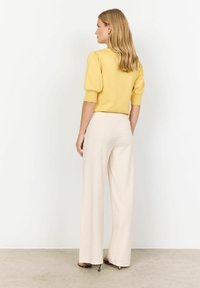 Haut en tricot jaune avec des manches bouffantes, associé à un pantalon large beige. Le modèle montre une vue de dos, avec un sol clair et un fond neutre.