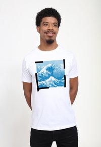 Hvid bomulds T-shirt med et blåt bølgemønster indrammet af sorte firkanter. Designet indeholder detaljerede bølgemønstre og indviklede detaljer.