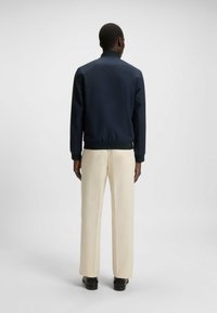 Navy bomber jacket with ribbed cuffs and waistband, paired with cream trousers and black shoes, viewed from the back.