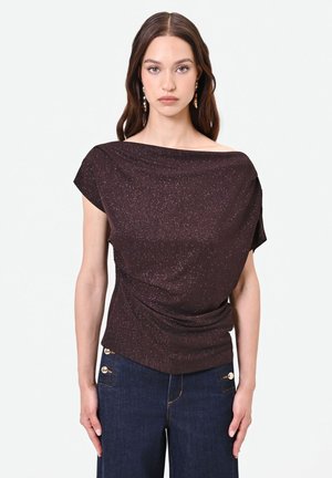 Donna con capelli lunghi e castani che indossa un top viola scuro glitterato con scollo a barca e jeans blu scuro con bottoni dorati, in piedi davanti a uno sfondo semplice.