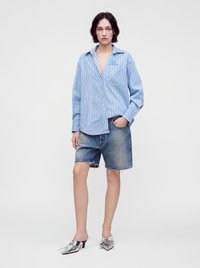 Γυναίκα όρθια, με μπλε και λευκό ριγέ oversized πουκάμισο, τζιν σορτς με κομμένα τελειώματα και ασημένια μυτερά mules με χαμηλό τακούνι σε λευκό φόντο.