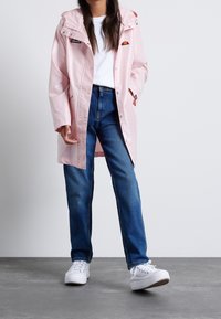 Osoba w jasnoróżowym płaszczu przeciwdeszczowym, białej koszulce, niebieskich dżinsach i białych trampkach stoi na szarym podłożu na tle białej ściany.