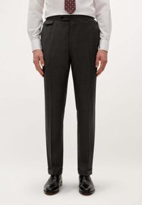 Hombre vestido con pantalones de traje gris oscuro, camisa blanca, corbata roja con estampado, y zapatos de vestir negros, de pie contra un fondo blanco.