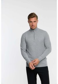 Pull à zip gris en matériau doux. Doté d'un col et de poignets côtelés, d'une coupe ajustée et d'une texture subtile. Pantalon noir visible.