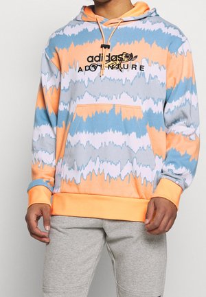 Osoba nosící vícebarevnou batikovanou mikinu v oranžové, modré a bílé barvě s nápisem "adidas adventure" a šedé tepláky.