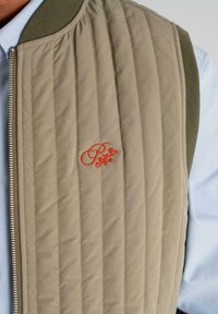 Beige quilted vest with vertical stitching and orange embroidered floral logo, worn over a light blue collared shirt.