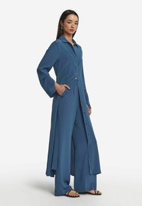 Lång blå klänning med kragkrage, knappstängd front, långa ärmar och sidofickor. Har en flödande design och avslappnad passform.