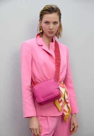 Bolso bandolera de cuero rosa con forma cuadrada, superficie texturizada y detalle de logo. Combinado con una blazer rosa y una bufanda colorida con estampado.