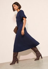 Marineblaues, strukturiertes Midikleid mit kurzen Ärmeln und gerafftem Oberteil, kombiniert mit kniehohen braunen Stiefeln mit Absatz und einer braunen Clutch.