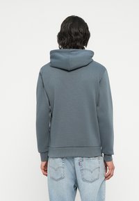 Felpa grigia con cappuccio, dal taglio rilassato, con polsini e orlo a coste. La parte posteriore presenta una texture liscia e dettagli di cucitura sottili.