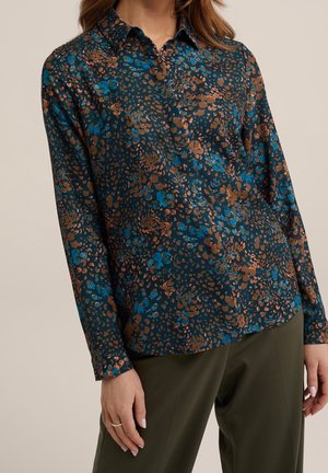 Vrouw draagt een blouse met lange mouwen en kraag, met een blauw-bruwn abstract bloemmotief, gecombineerd met olijfgroene broek.