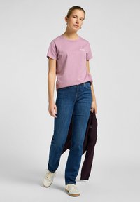 Camiseta de algodón rosa con el texto "PAZ", combinada con jeans de pierna recta azul oscuro y zapatillas blancas. El modelo sostiene una chaqueta oscura.