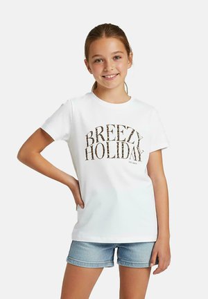 Meisje met lichtbruin haar in een paardenstaart, draagt een wit t-shirt met de tekst "Breezy Holiday" en blauwe spijkershorts, glimlacht met één hand op haar heup.