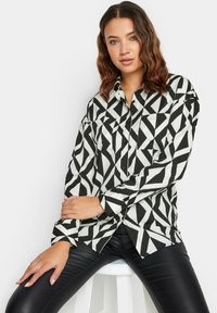 Camisa de botones en blanco y negro con patrón geométrico, que presenta un cuello recto, mangas largas y dos bolsillos en el pecho.
